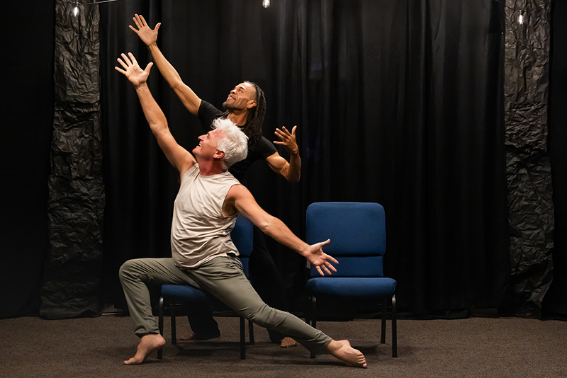 Octavio Campos and Darryl Brown pose dynamically on a small stage between blue chairs, extending their arms upward and outward against a backdrop of black curtains.