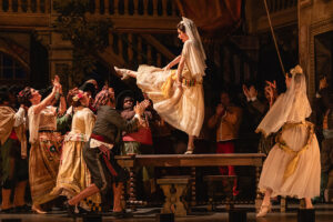 A costumed stage ensemble performs a lively theatrical dance scene on a warmly lit set, with Sydney balanced atop a table while surrounding performers reach upward amid an ornate interior backdrop of staircases and balconies.