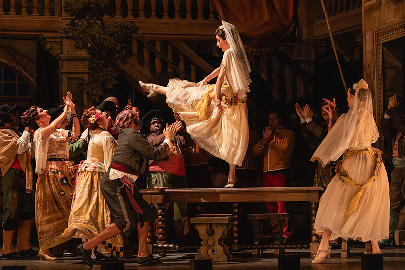 A costumed stage ensemble performs a lively theatrical dance scene on a warmly lit set, with Sydney balanced atop a table while surrounding performers reach upward amid an ornate interior backdrop of staircases and balconies.