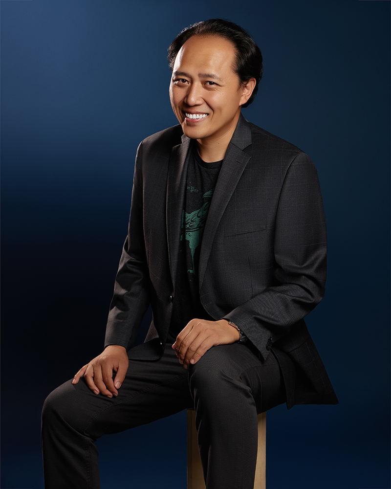 : Haowen is a Taiwanese man with dark, wavy hair, warm brown eyes, and a light-medium complexion. Wearing a charcoal suit over a dark shirt, he sits on a stool with relaxed posture, hands resting naturally. He smiles openly toward the camera against a smooth deep-blue backdrop.