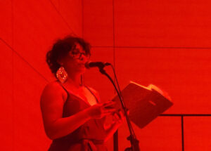 Amber stands at a microphone under intense red lighting, reading from an open book during a live poetry or spoken-word presentation.
