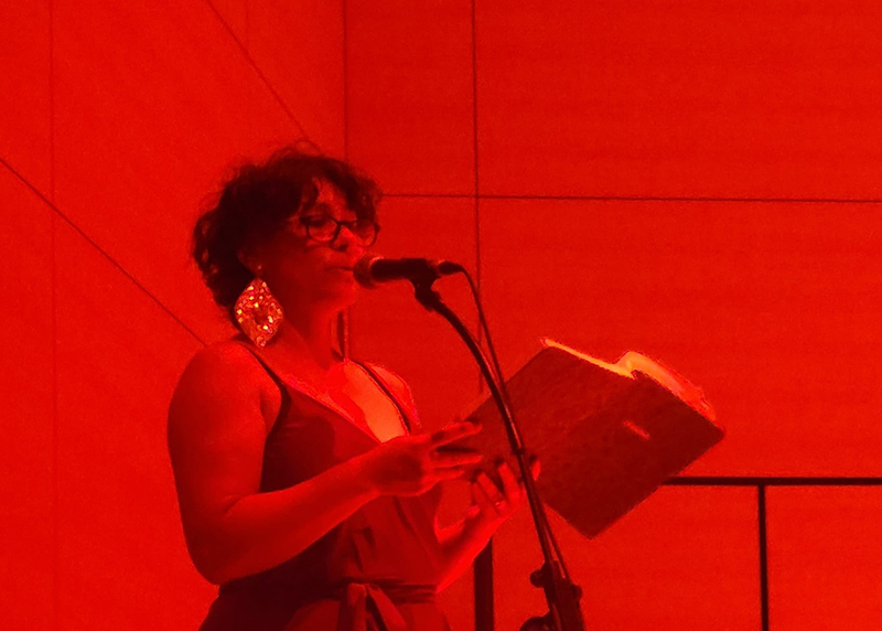 Amber stands at a microphone under intense red lighting, reading from an open book during a live poetry or spoken-word presentation.