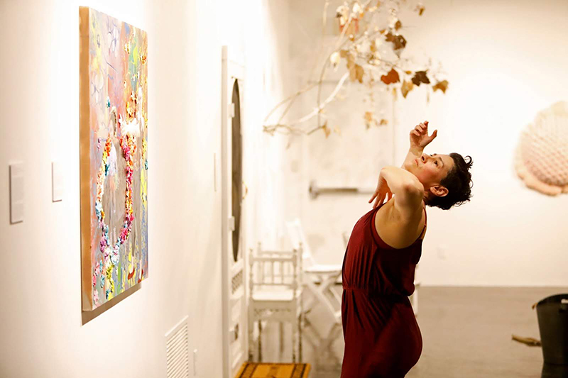Amber, wearing burgundy, dances with uplifted arms and face beside a colorful wall-mounted artwork in a softly lit gallery space featuring sculptural installations and white walls.
