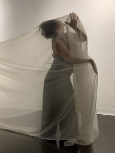 Amber, dressed in dark clothing, stands enveloped in flowing translucent fabric, creating a sculptural silhouette against a minimalist gallery backdrop.