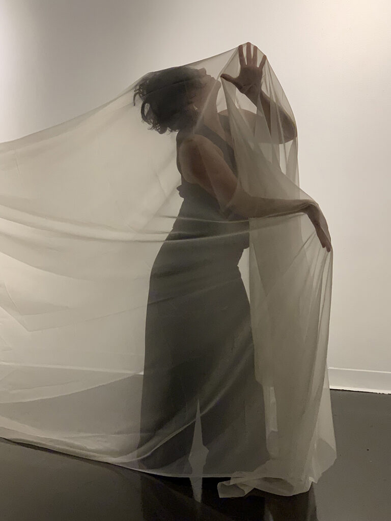 Amber, dressed in dark clothing, stands enveloped in flowing translucent fabric, creating a sculptural silhouette against a minimalist gallery backdrop.