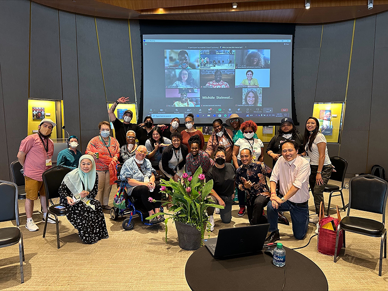 A group photo of the Round 2 DFA Artist Fellows and program team. Twenty or so people pose together, some kneeling on the ground, some in chairs, and some standing. A large screen appears behind them, with photos representing the Fellows who are joining the group virtually. Photo by Michele Steinwald.