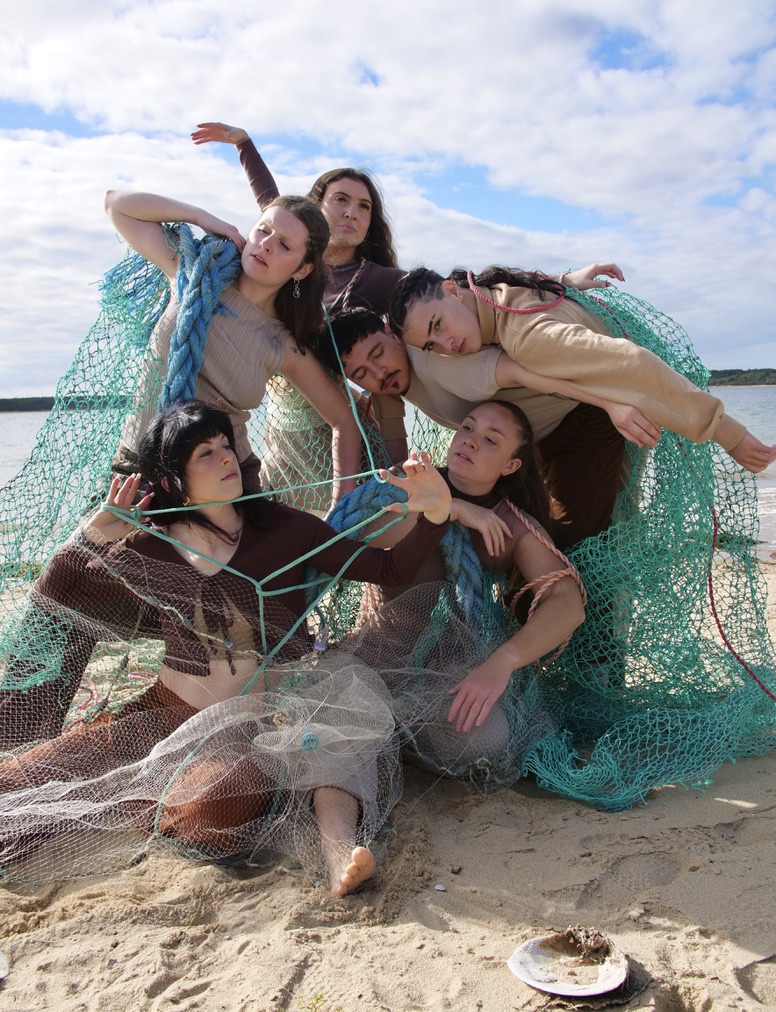Five performers pose together on a sandy shoreline, draped and entwined in fishing nets and rope, creating a sculptural, interconnected composition against a backdrop of water and a cloudy sky.