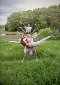 A group of performers in a grassy field creates a layered, tree-like formation with outstretched arms and balanced poses, forming a striking collaborative sculpture against a backdrop of lush greenery and an overcast sky.