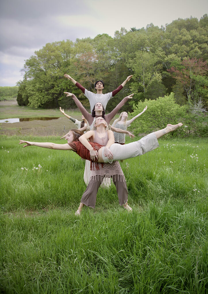 A group of performers in a grassy field creates a layered, tree-like formation with outstretched arms and balanced poses, forming a striking collaborative sculpture against a backdrop of lush greenery and an overcast sky.