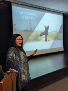Katie stands beside a podium while giving a talk in an auditorium, with a large projection screen behind displaying the title “LIQUID SPINE, AUGUSTA” and an image of a dancer in an outdoor industrial landscape.