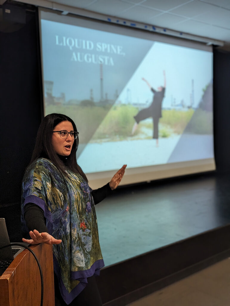 Katie stands beside a podium while giving a talk in an auditorium, with a large projection screen behind displaying the title “LIQUID SPINE, AUGUSTA” and an image of a dancer in an outdoor industrial landscape.