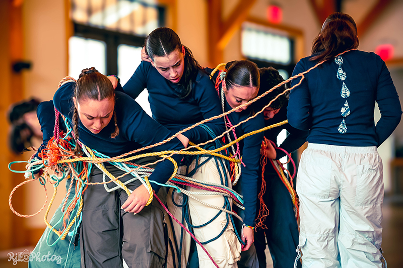 A group of dancers wearing dark tops and loose pants huddle together while entangled in a web of colorful ropes during a collaborative movement activity in an indoor hall.