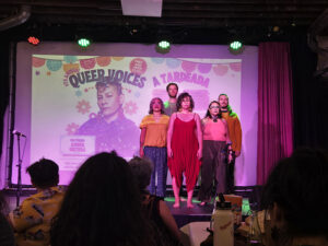 Five performers stand in a tight formation on a small stage before a projected event poster, presenting a collective live performance to an audience.