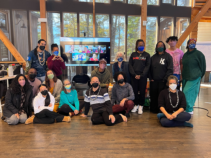 A group photo of the Round 3 DFA Artist Fellows and program team. Twenty or so people pose together, some sitting on the ground and some standing. A wall of windows and a large screen appears behind them, with photos representing the Fellows who are joining the group virtually. Photo courtesy of Dance/USA.