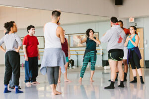 A group of dancers stands in a studio rehearsal circle as an instructor demonstrates movement and guides discussion in a collaborative workshop setting.
