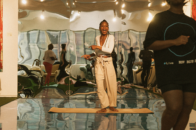 djassi stands barefoot on a mat in front of a large wavy mirrors, surrounded by distorted reflections and warm overhead lights in an indoor studio.