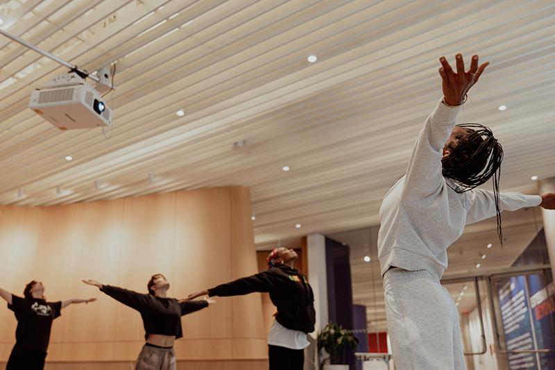 djassi leads four students in extending their arms upward and outward beneath a ceiling-mounted projector in a spacious indoor studio with warm lighting and wood-paneled walls.