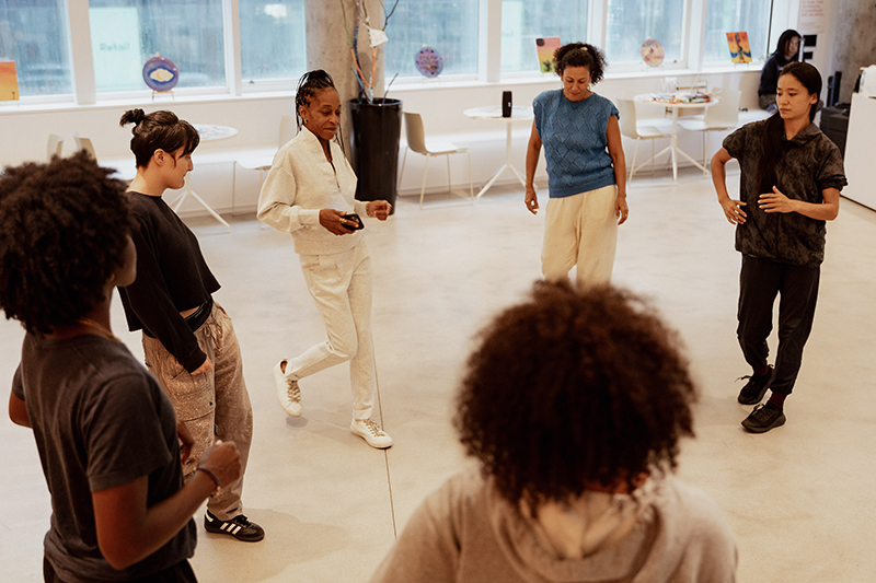 djassi, wearing all white, stands in a circle of other dancers in a bright studio-like space, practicing guided movement exercises on an open floor surrounded by tables, chairs, and large windows.
