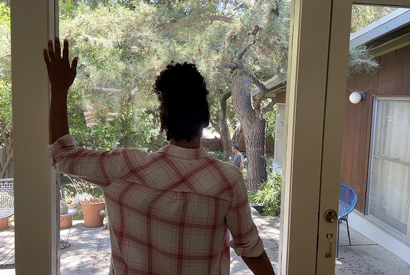 A person stands inside a doorway with one hand on the frame, looking out toward a sunlit patio and tree-filled courtyard framed by mid-century style buildings.