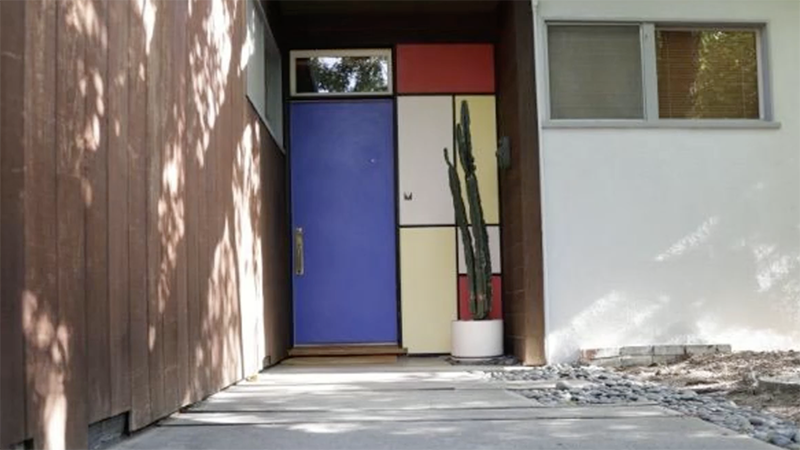 A colorful mid-century modern entryway features a bold blue door framed by geometric red, yellow, and white panels, with a tall potted cactus standing beside the entrance.