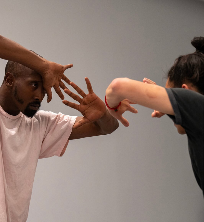Two performers face each other, extending and framing their hands around their faces against a plain background.