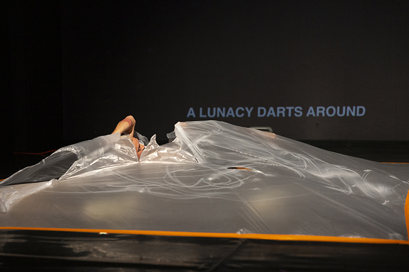 A performer lies partially covered beneath a large translucent plastic sheet on a dark stage, while the projected text “A LUNACY DARTS AROUND” appears in the background.