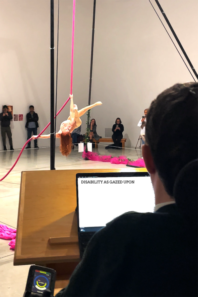 Erin with red hair flowing hangs inverted from bright pink aerial silk in a gallery-like performance space, observed by a lecturer seated in a wheelchair behind a laptop displaying the words “DISABILITY AS GAZED UPON.”
