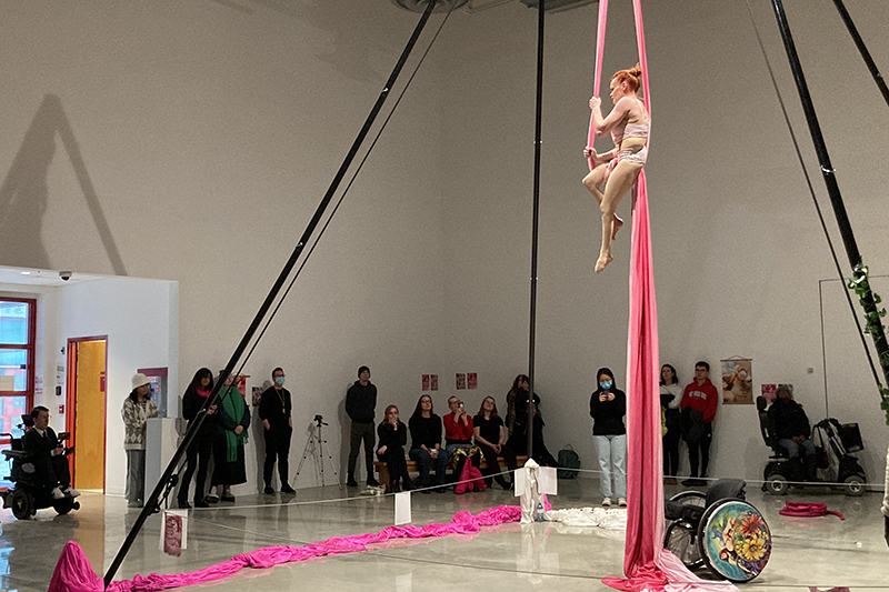 Erin sits in vivid pink silks suspended from a freestanding rig in a gallery space, while seated and standing audience members observe from around the performance area, where a decorated wheelchair is visible near the base of the apparatus.