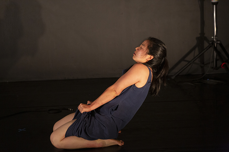 A performer in a sleeveless gray dress kneels on a dimly lit stage, leaning backward and clutching her dress fabric while stage equipment is faintly visible in the background.