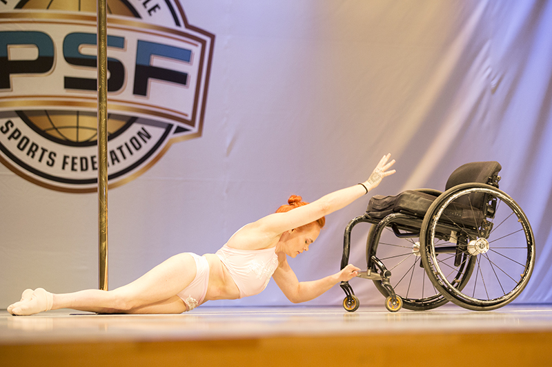 Erin stretches in a light two-piece costume across a stage toward her wheelchair and beside a pole, all in front of a large IPSF backdrop.