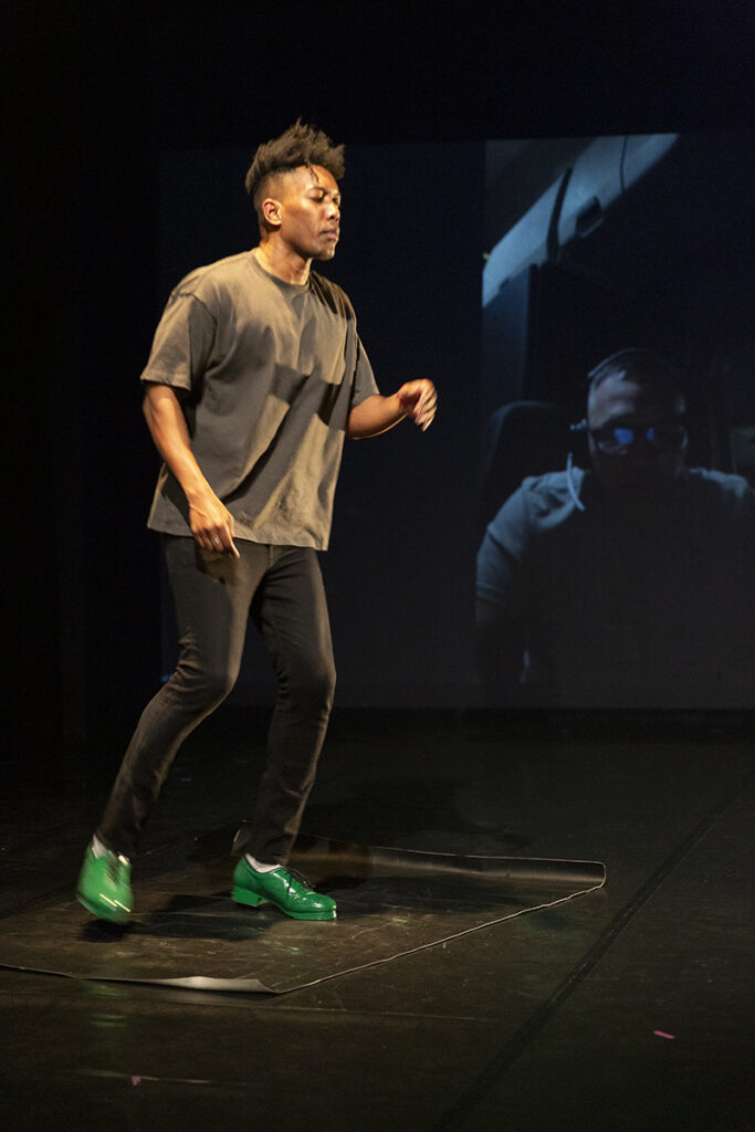 A tap dancer with bright green shoes performs on a dimly lit stage, with a projection of a video call participant visible in the background.