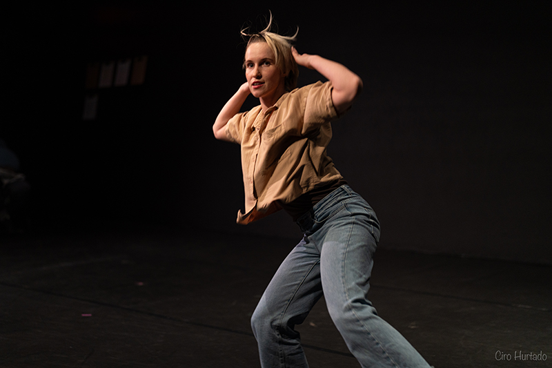 A performer in a beige shirt and blue jeans lifts her hands to her head with bent knees on a dimly lit stage, her hair lifted in motion under theatrical lighting.