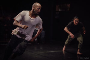Two performers run across a dimly lit stage, one wearing a white shirt and the other black.