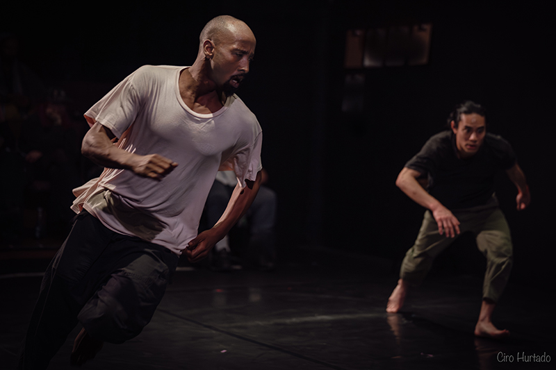 Two performers run across a dimly lit stage, one wearing a white shirt and the other black.